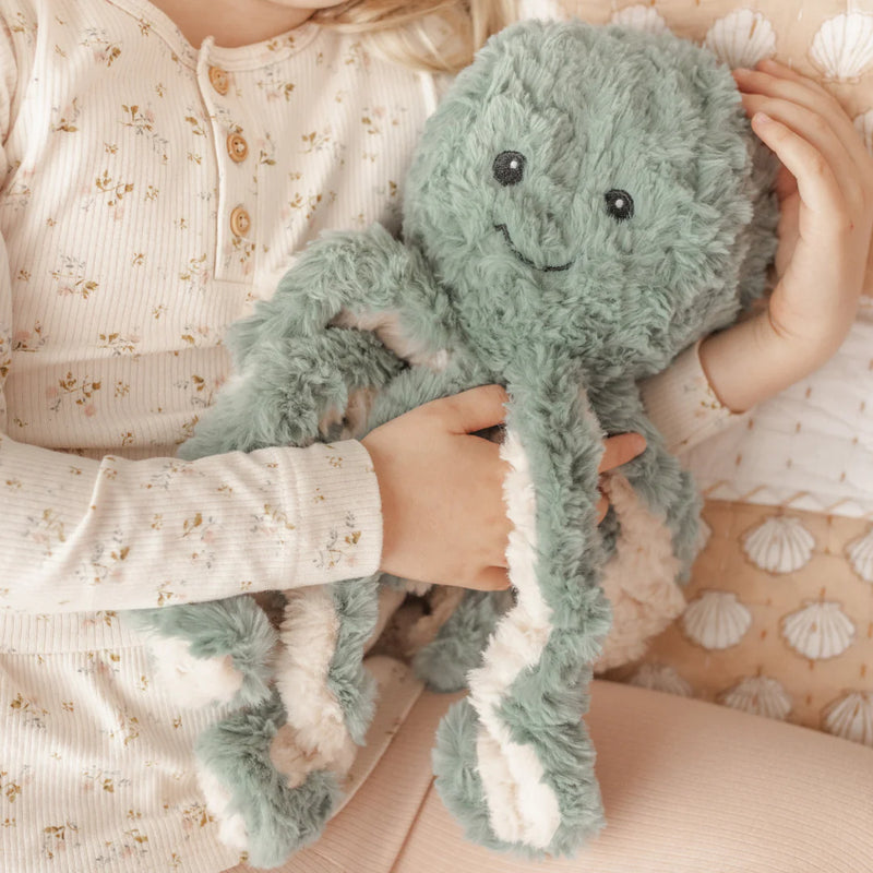 Child holding a sage green octopus shaped weighted toy on patterned bedding