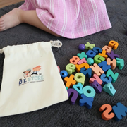 Colourful alphabet lacing beads scattered beside a branded drawstring bag on a dark carpet, next to a child wearing a pink striped dress.