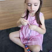 Young child in pink dress sitting on a rug threading colourful beads onto a blue string for fine motor play.