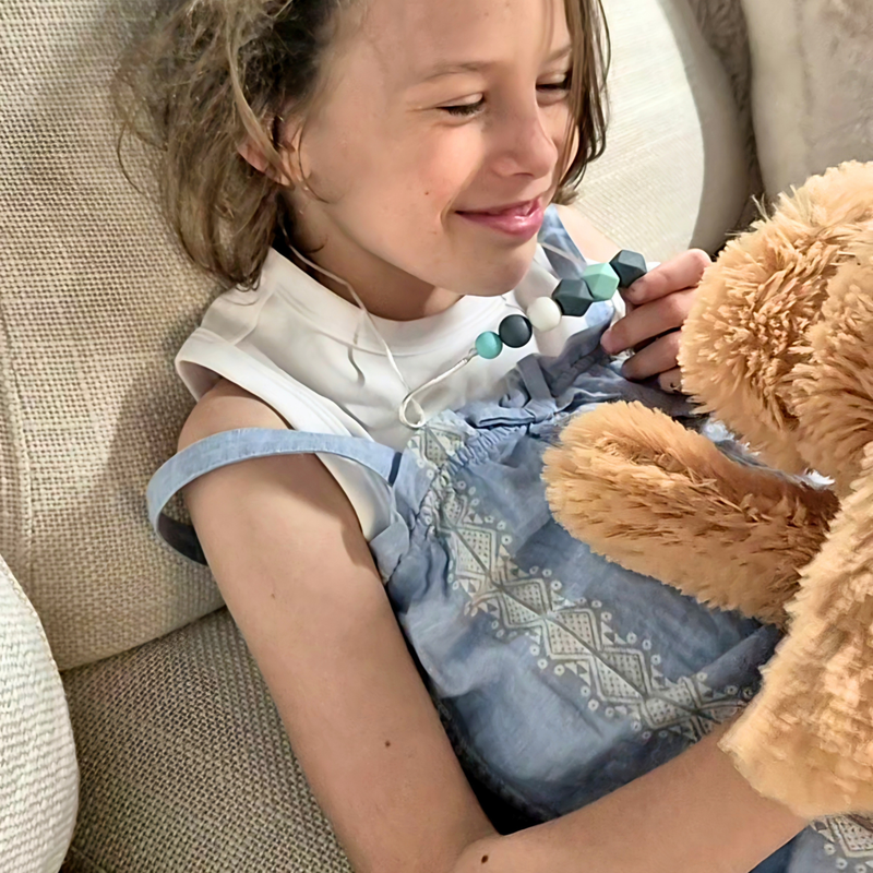 Child wearing a blue and grey beaded chew pendant necklace while cuddling a teddy bear on a sofa