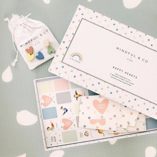 Open board game box with pastel polka dot design, heart-shaped cards, and a small drawstring bag holding colorful wooden game pieces.