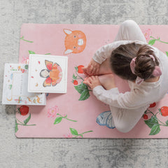 Child sitting cross-legged on a pink kids yoga mat with sweet print featuring animals and strawberries, next to an illustrated activity book.