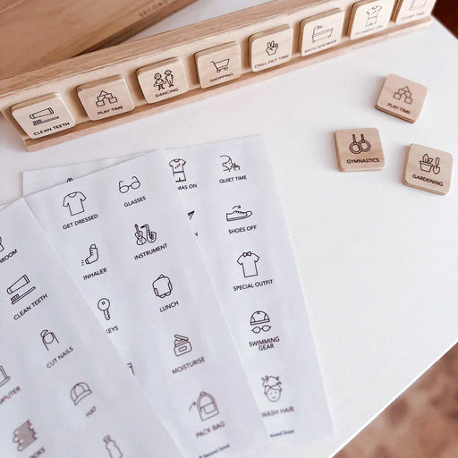 Wooden routine and reminder labels with simple black icons showing daily activities, alongside printed sheets with matching icons on a white surface.